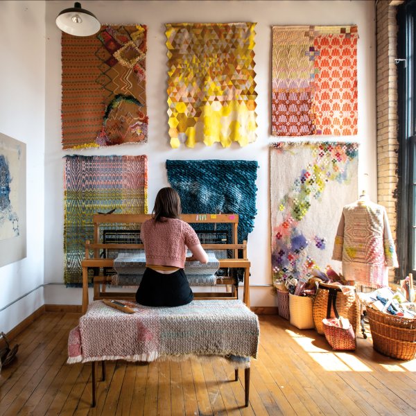 Jensen at her loom weaving cloth for a series of new artworks. To her right are baskets of cloth and a wearable woven coat. On the wall are her woven, quilted, and embroidered pieces.