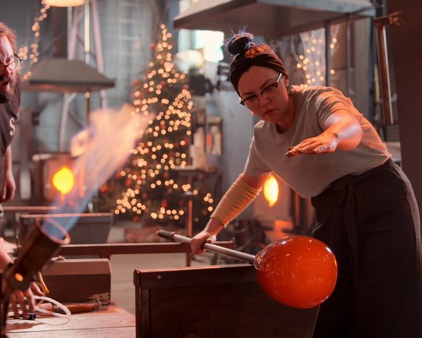 Glass blower works on a vessel in studio with christmas tree in the background