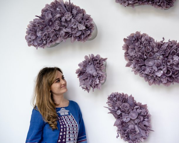 portrait of artist looking up at a series o wall sculptures that look like bunches of flower pedals made from purple glass all hanging on a white wall