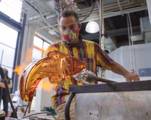 glass artists working an orange arrow-shaped glass sculpture with flame