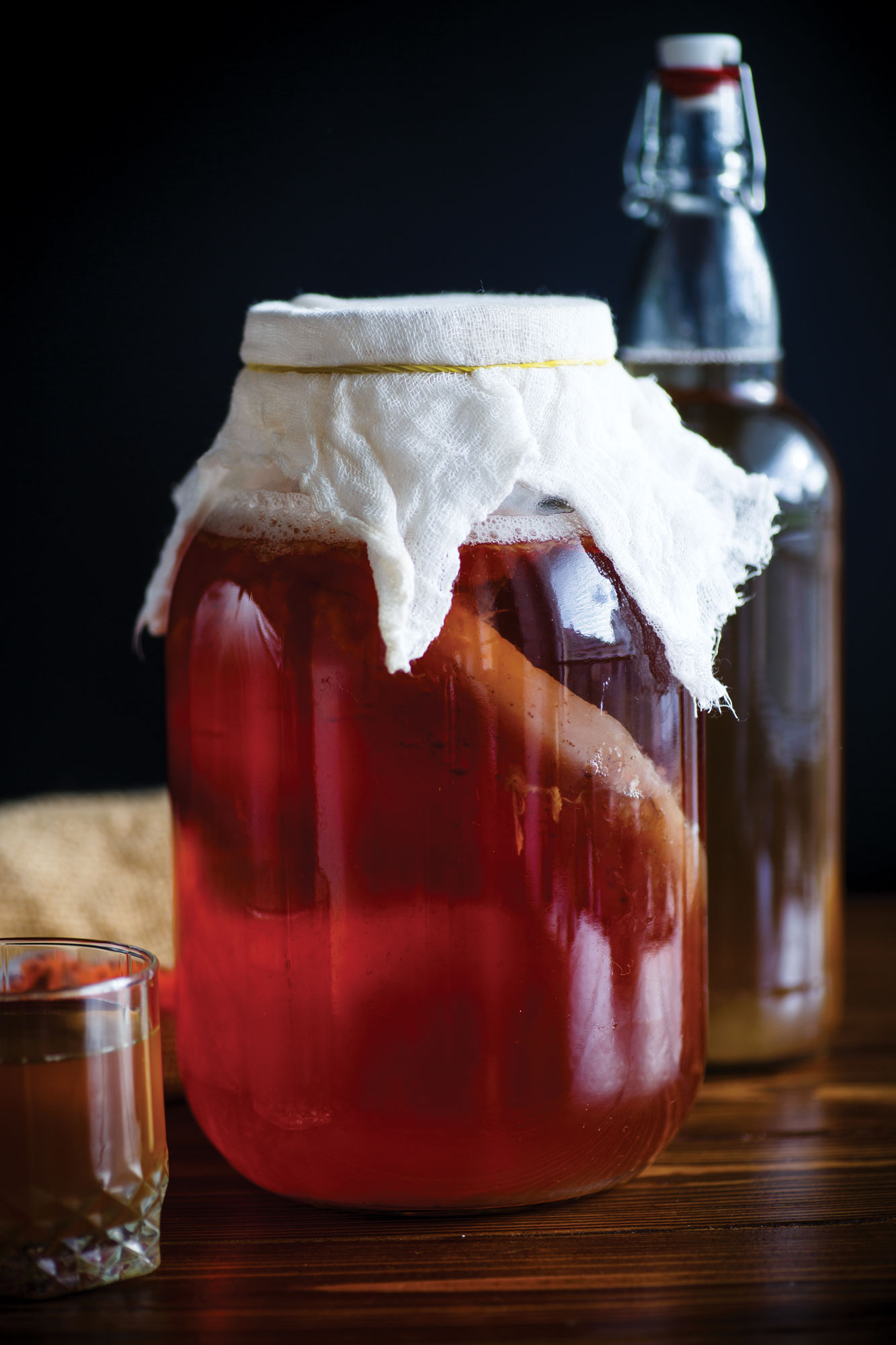 A jar of kombucha fermenting with a bottle in the background