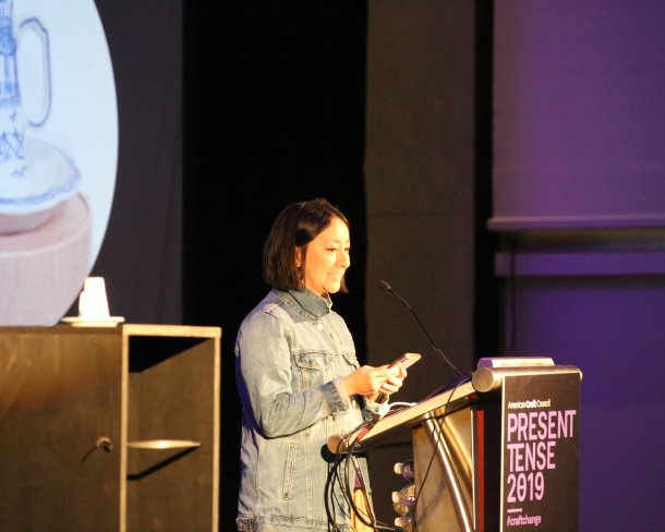 Person presenting at a podium beside a ceramic cup on a pedestal with slideshow image in backdrop