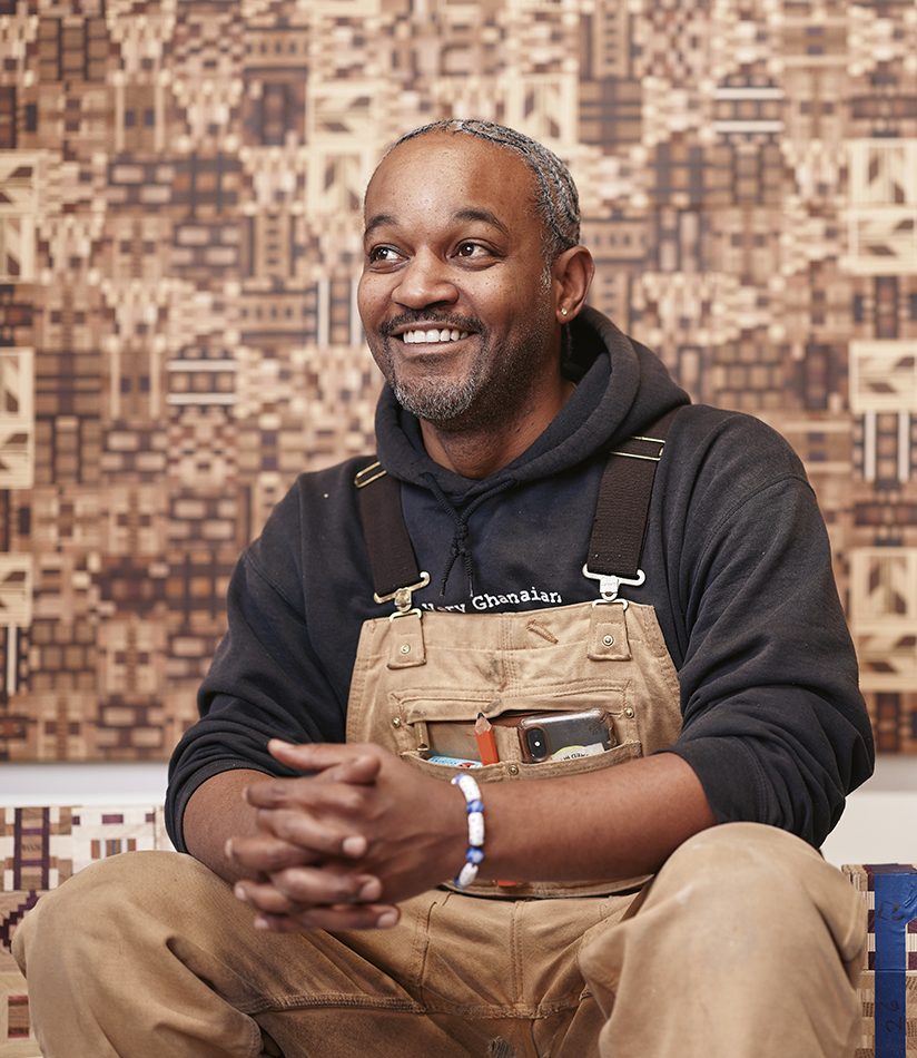 Ato Ribeiro in his studio, wearing overalls over a sweatshirt, with artwork behind him.