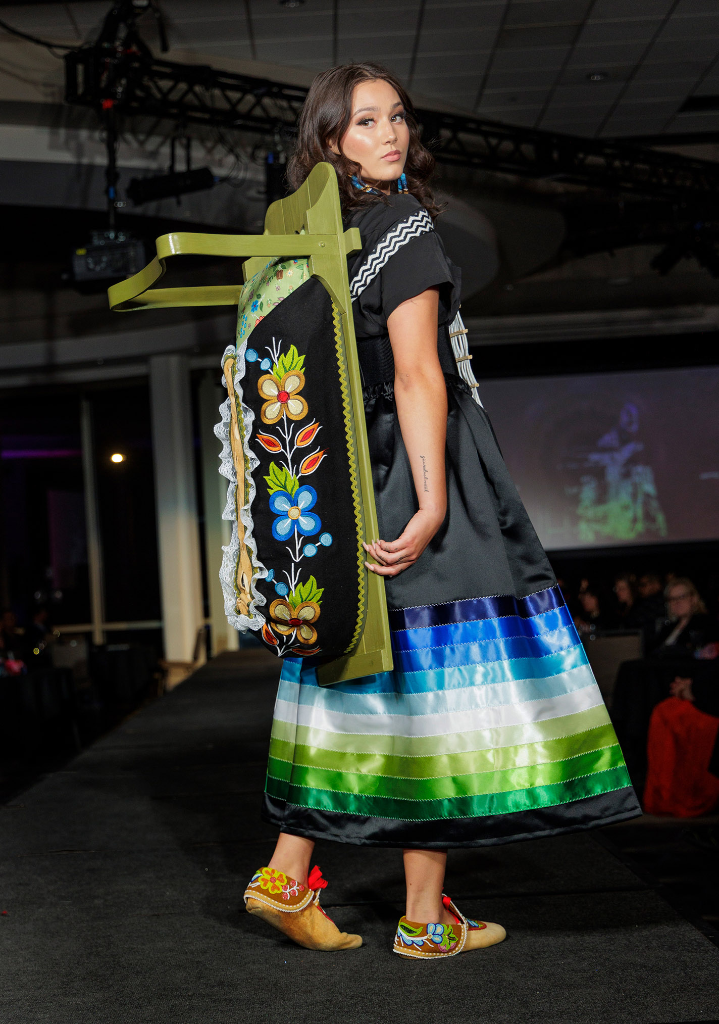 Model wears a look featuring an interpretation of a traditional ribbon skirt plus an intricately beaded traditional cradleboard.